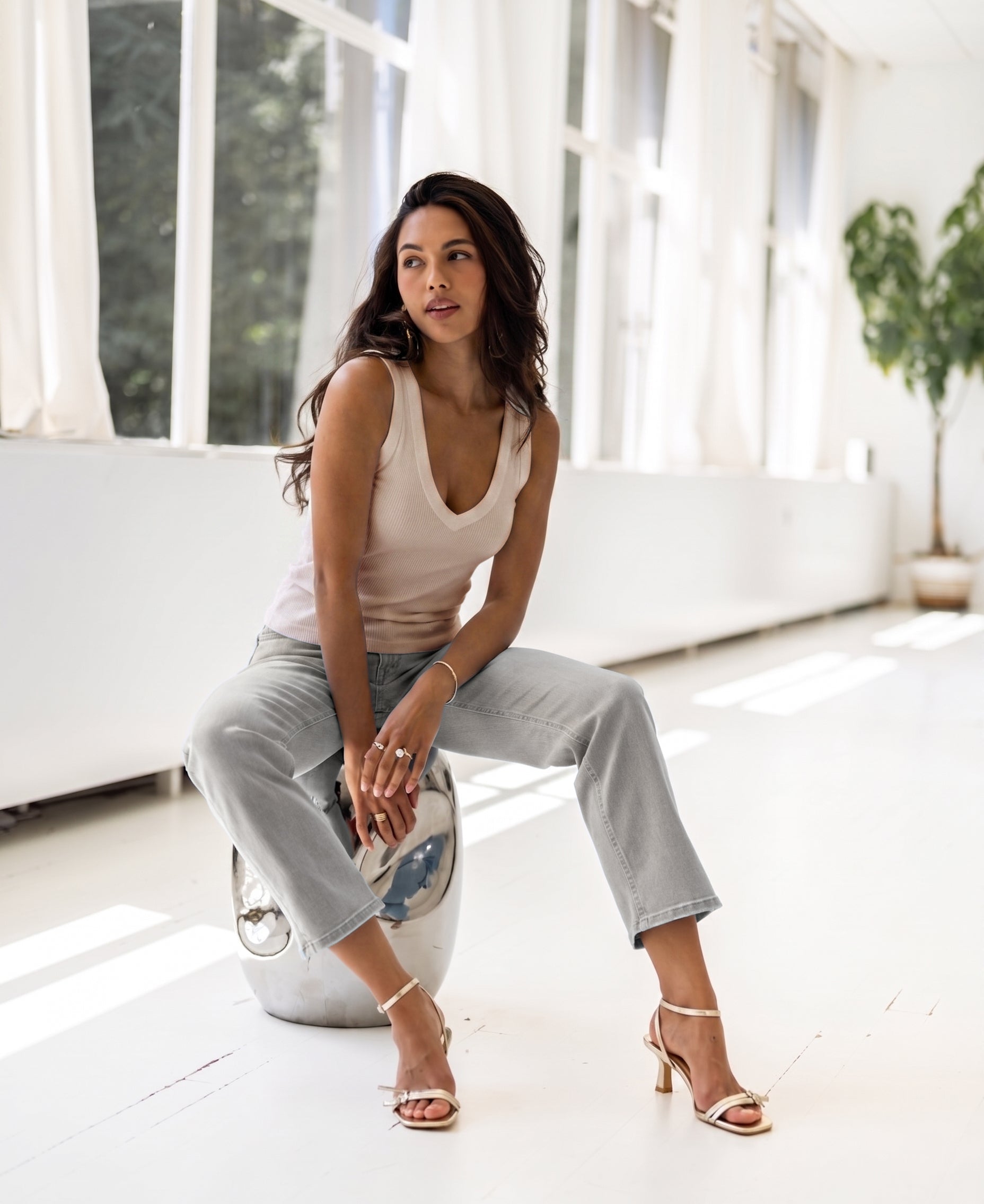 Woman wearing a luxe and soft butter-colored top sits on a modern stool in a bright, plant-filled room.