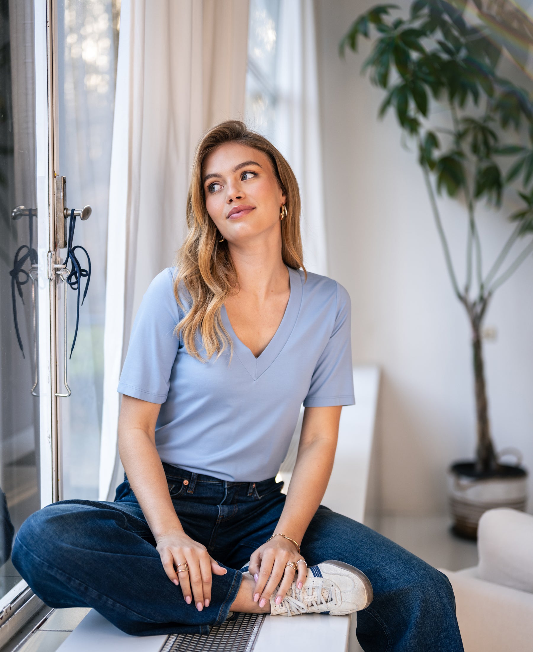 Light blue LA VERONIQUE V-neck T-shirt by PLEIN PUBLIQUE shown worn by a woman sitting cross-legged.