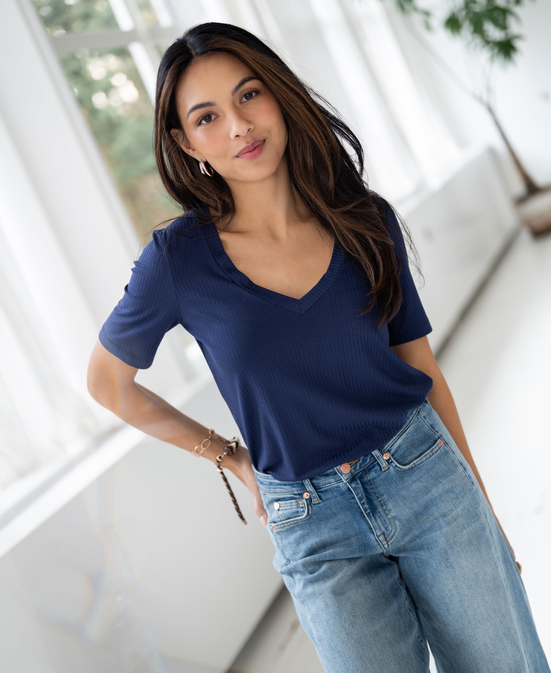 Woman wears the PLEIN PUBLIQUE Bamboo T-shirt LES VERSAILLES in indigo, inside, hand in pocket, smiling gently.