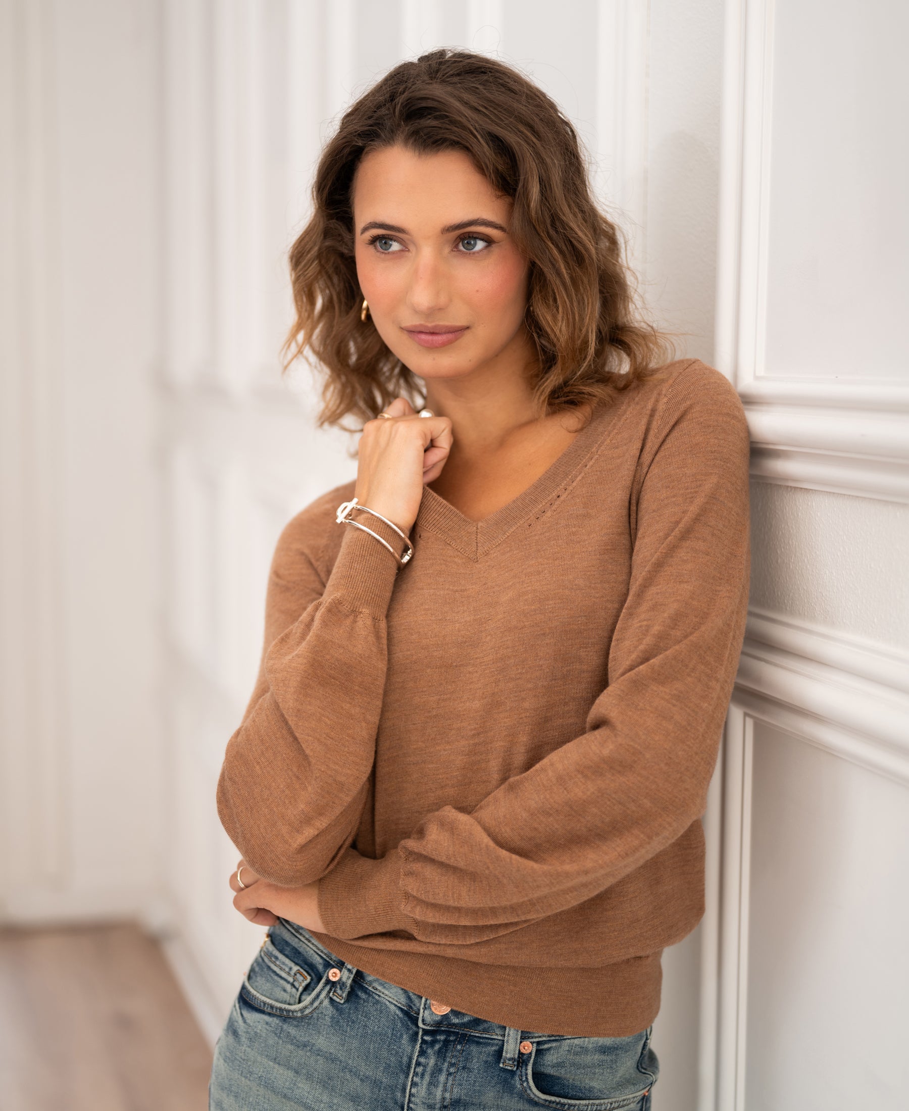 Woman wearing a PLEIN PUBLIQUE Merino wool V-neck sweater in Chai tea color stands by a white wall.