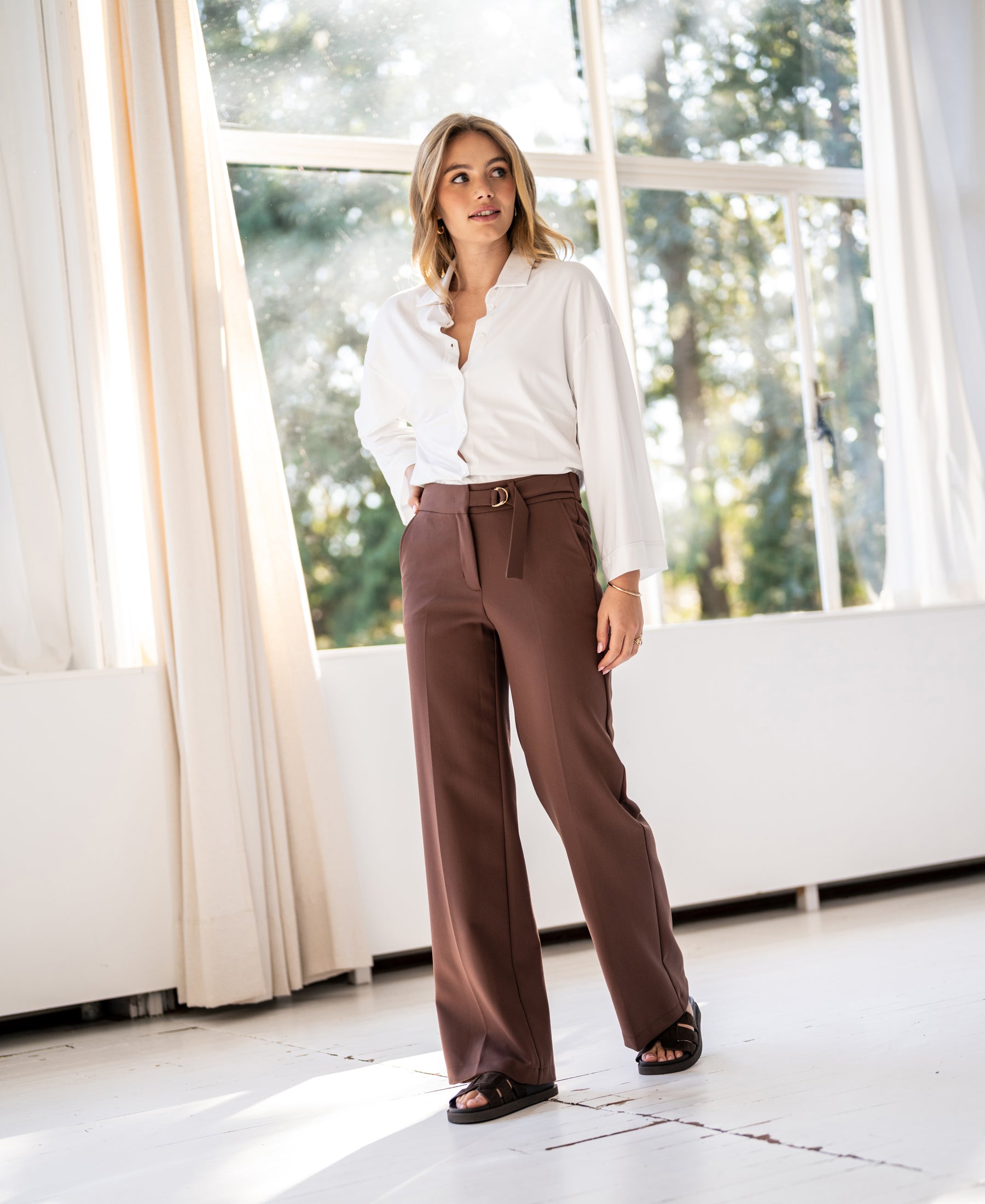 Woman wears brown LA WENDE pants by PLEIN PUBLIQUE, standing indoors near sunlit windows.