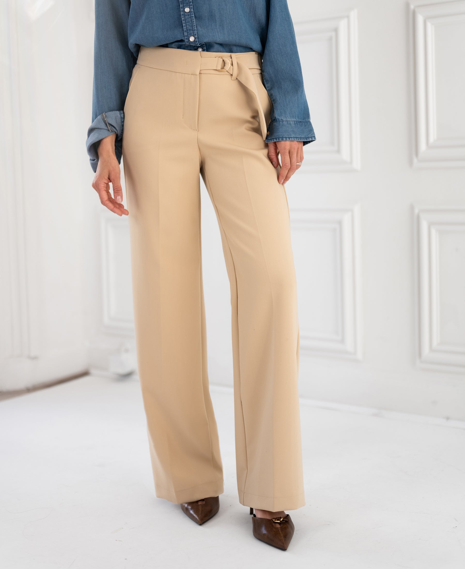 A person wears PLEIN PUBLIQUE’s LA WENDE Sand Trousers, standing on a white floor.