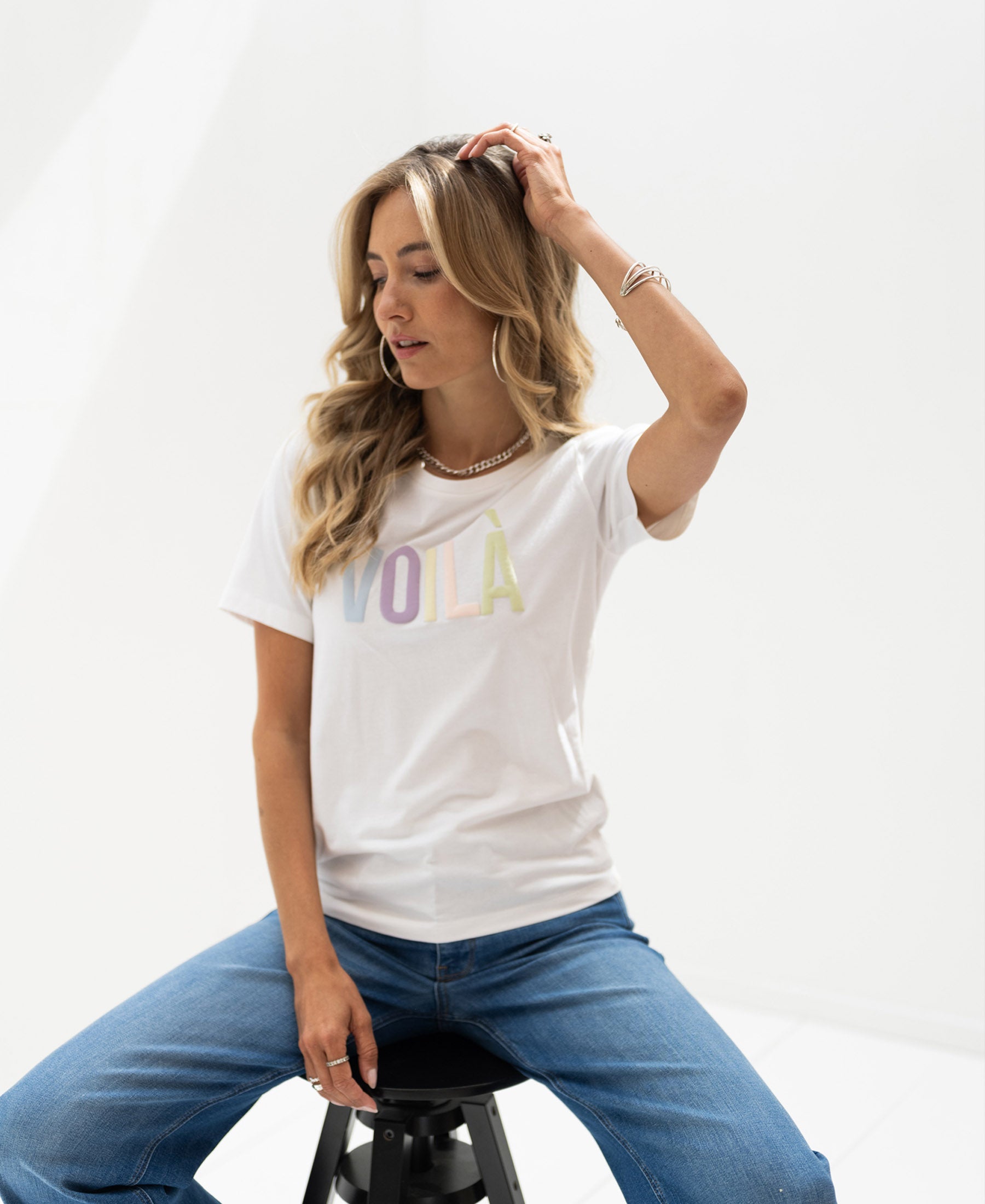 Woman wearing a white cotton Voila t-shirt sits on a stool, touching her hair.
