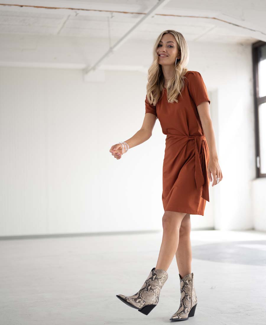 Woman wearing the Plein Publique Button Dress in terra color, smiling in a bright, minimalist room.
