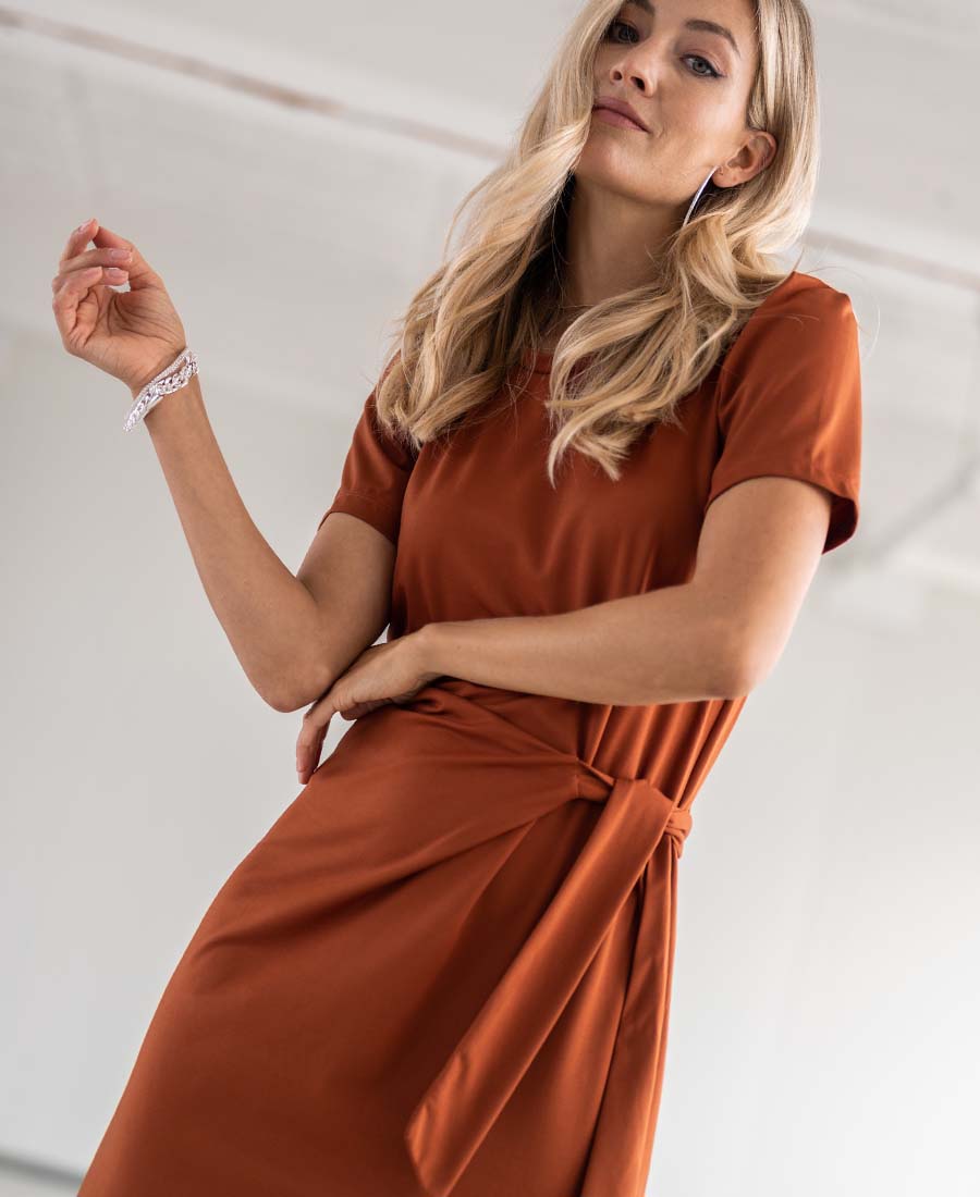A woman models the PLEIN PUBLIQUE tie-front dress LA FEE in Terra color indoors.