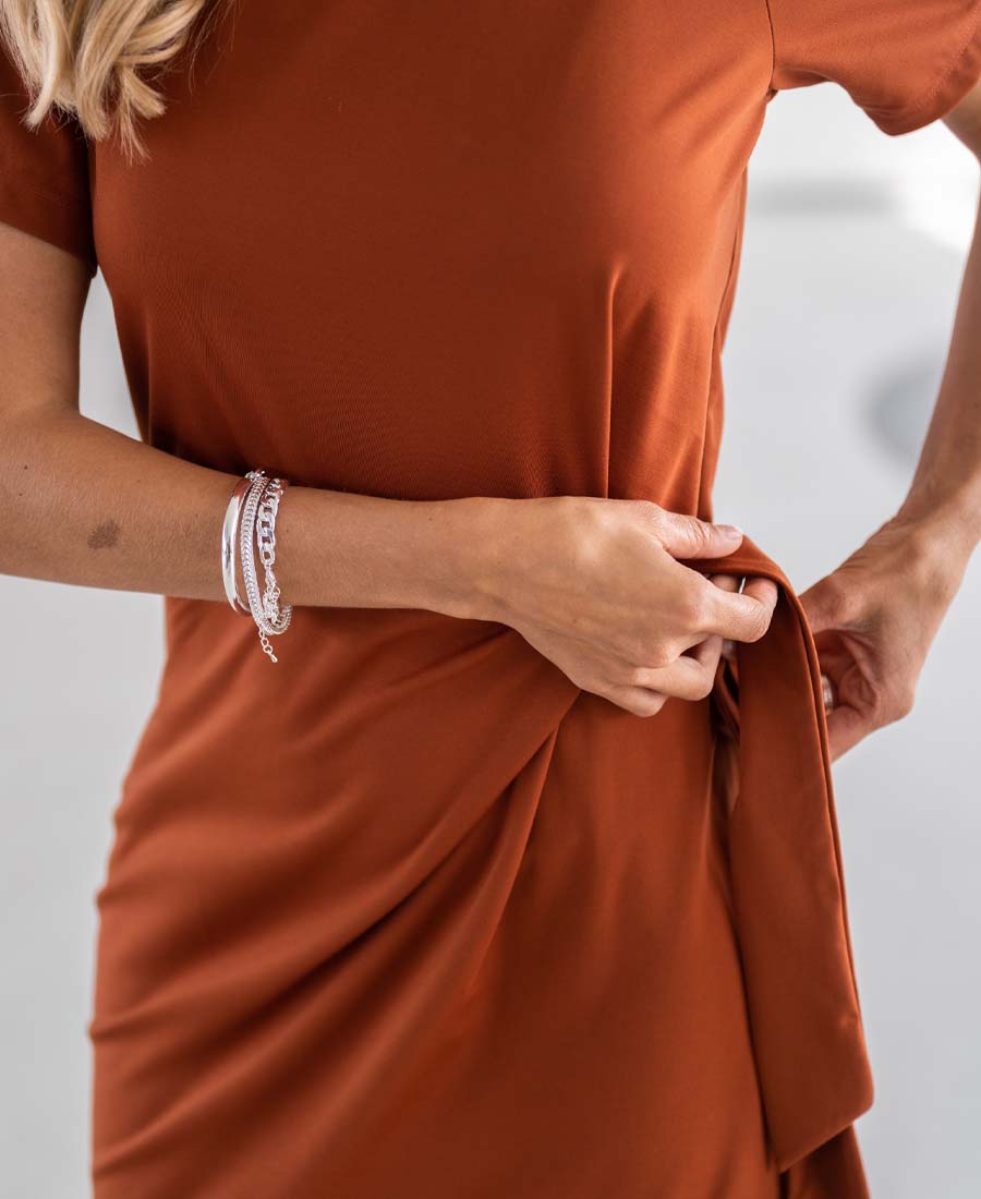 Woman wearing the Terra Button Dress in brown by PLEIN PUBLIQUE, tying a side knot indoors.