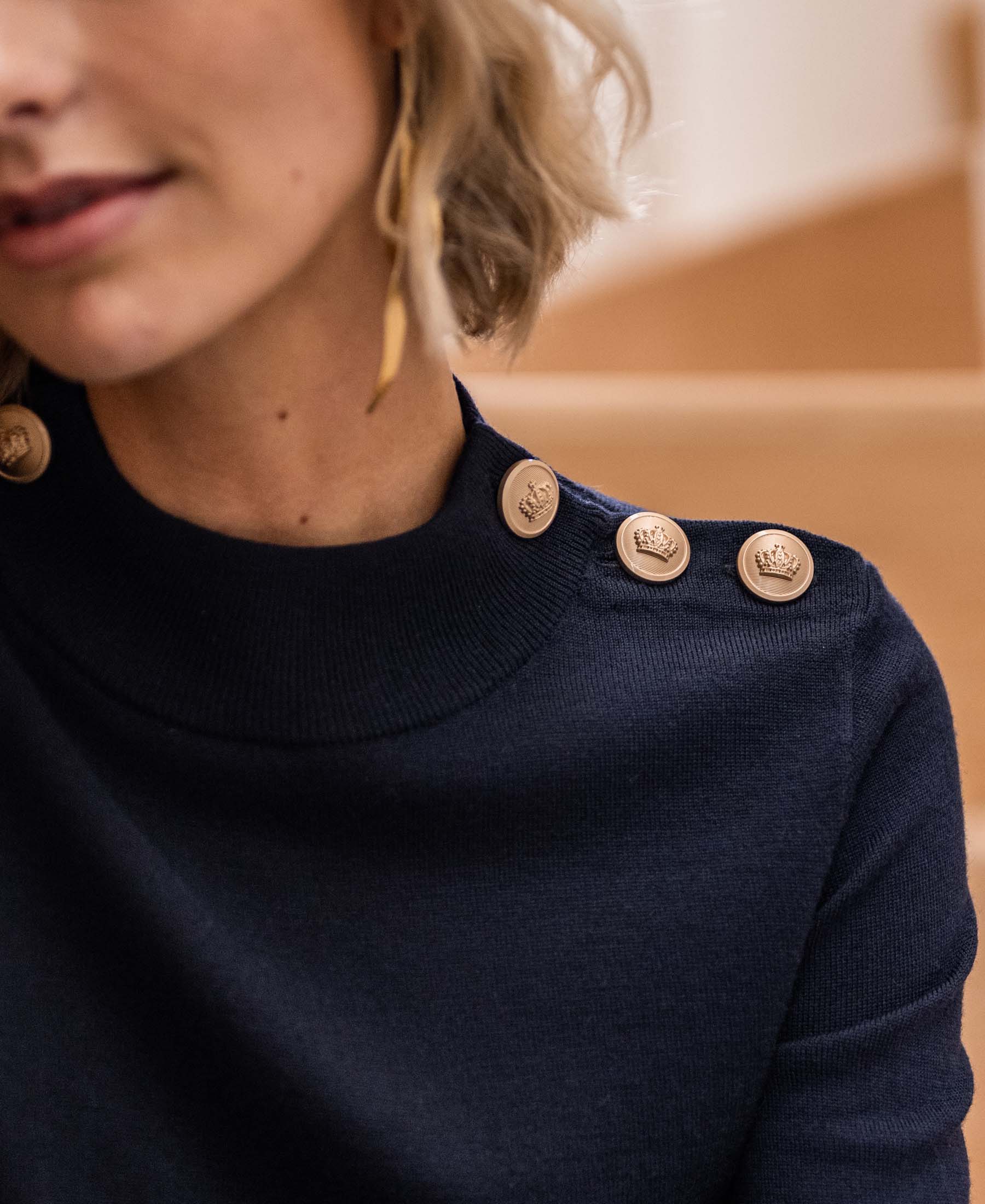 Navy blue merino wool sweater with gold buttons, partial front view. Brand name visible; model's face partly shown.