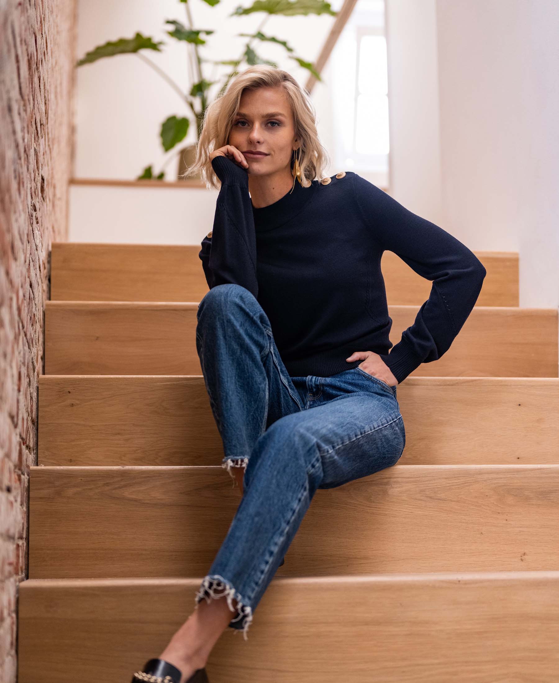 Woman wearing a navy blue Merino sweater by PLEIN PUBLIQUE sits on wooden stairs. Plant in background.