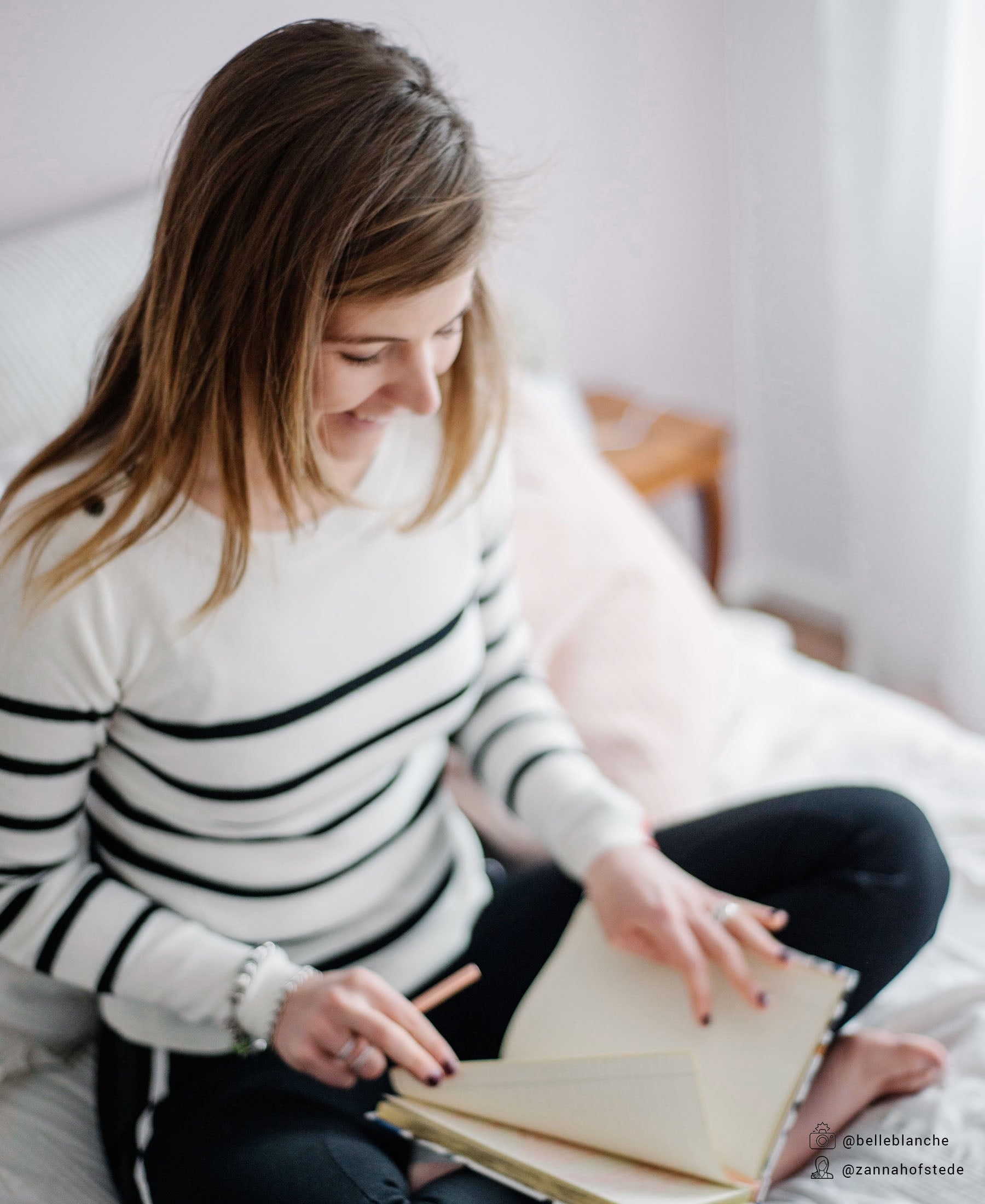 Woman wearing an ivory-black Breton striped sweater sits on a bed, smiling as she writes in a notebook.