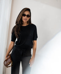 Black heart-patterned sweater by PLEIN PUBLIQUE, shown worn by a woman standing by a bright window.
