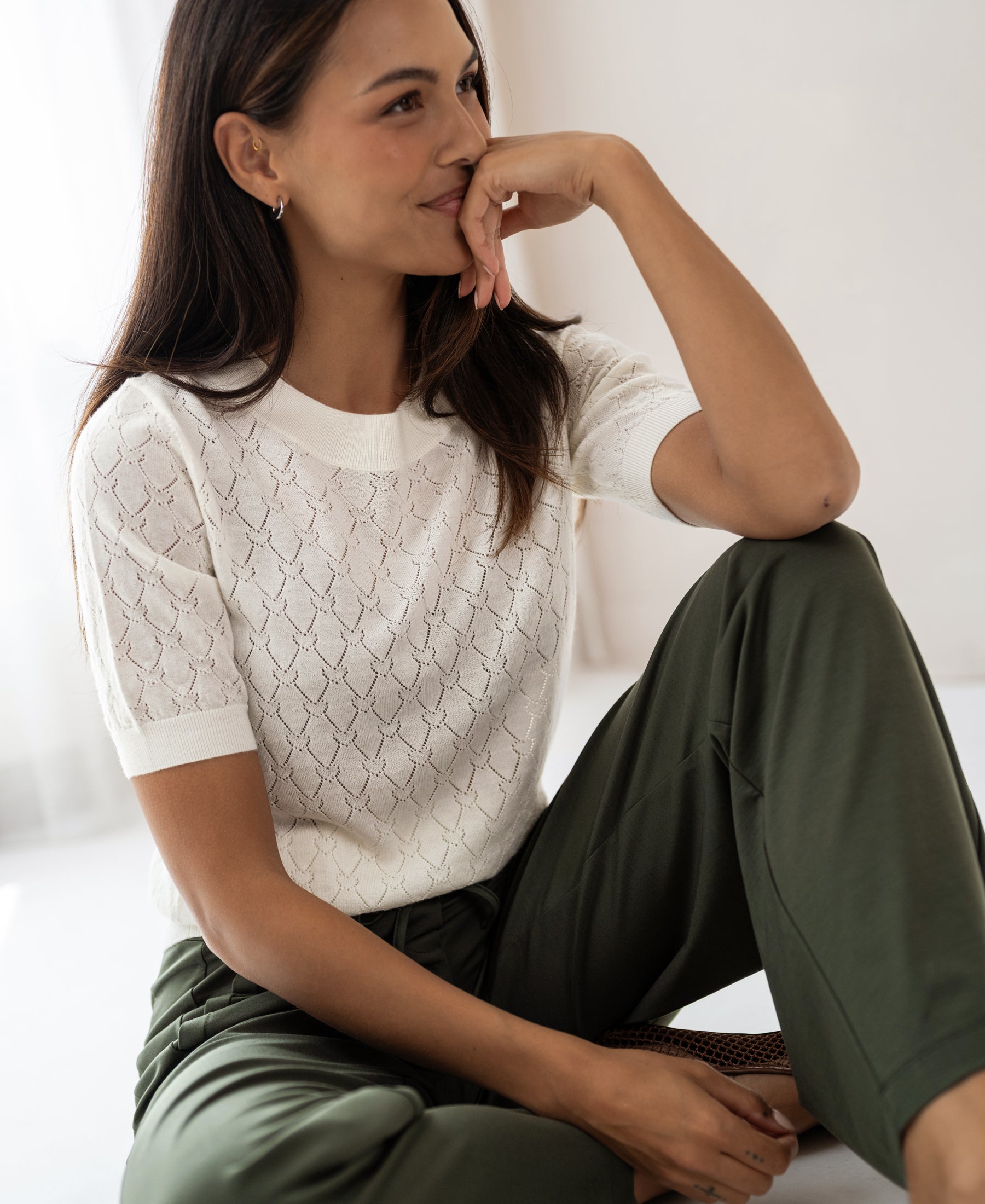 Woman wearing an ivory sweater with heart pattern from PLEIN PUBLIQUE LE BATTANT sits smiling on the floor in a bright room.