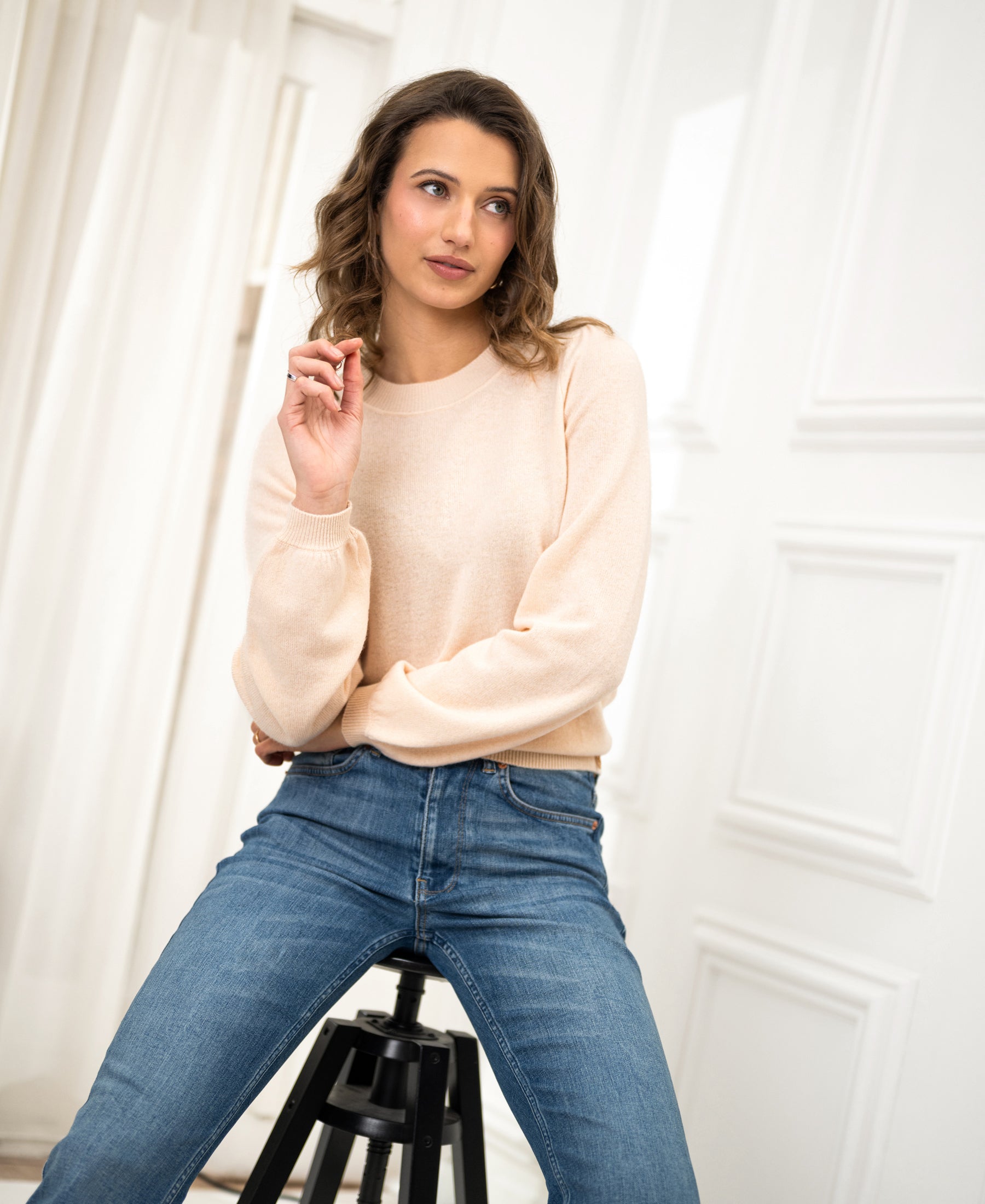 Woman wearing the LA BAYONNE Butter merino sweater by PLEIN PUBLIQUE, seated on a stool in a bright room.
