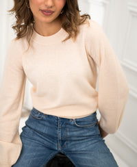 Woman wearing a PLEIN PUBLIQUE LA BAYONNE butter-colored merino wool sweater sits on a stool in a bright room.