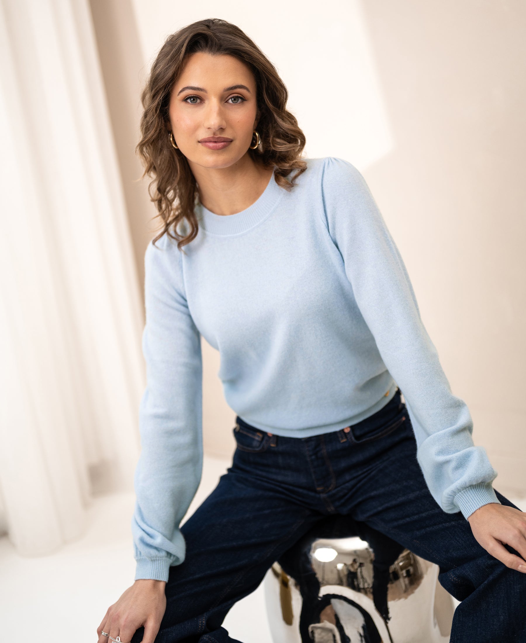 Soft merino wool sweater LA BAYONNE in light blue from PLEIN PUBLIQUE, shown worn by a woman seated on a silver stool.