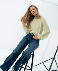 Woman wearing a soft merino wool sweater in light green, seated on a stool in a bright studio.