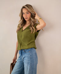 Sleeveless olive green blouse by PLEIN PUBLIQUE, worn by a woman with long wavy blonde hair, standing by a beige wall.