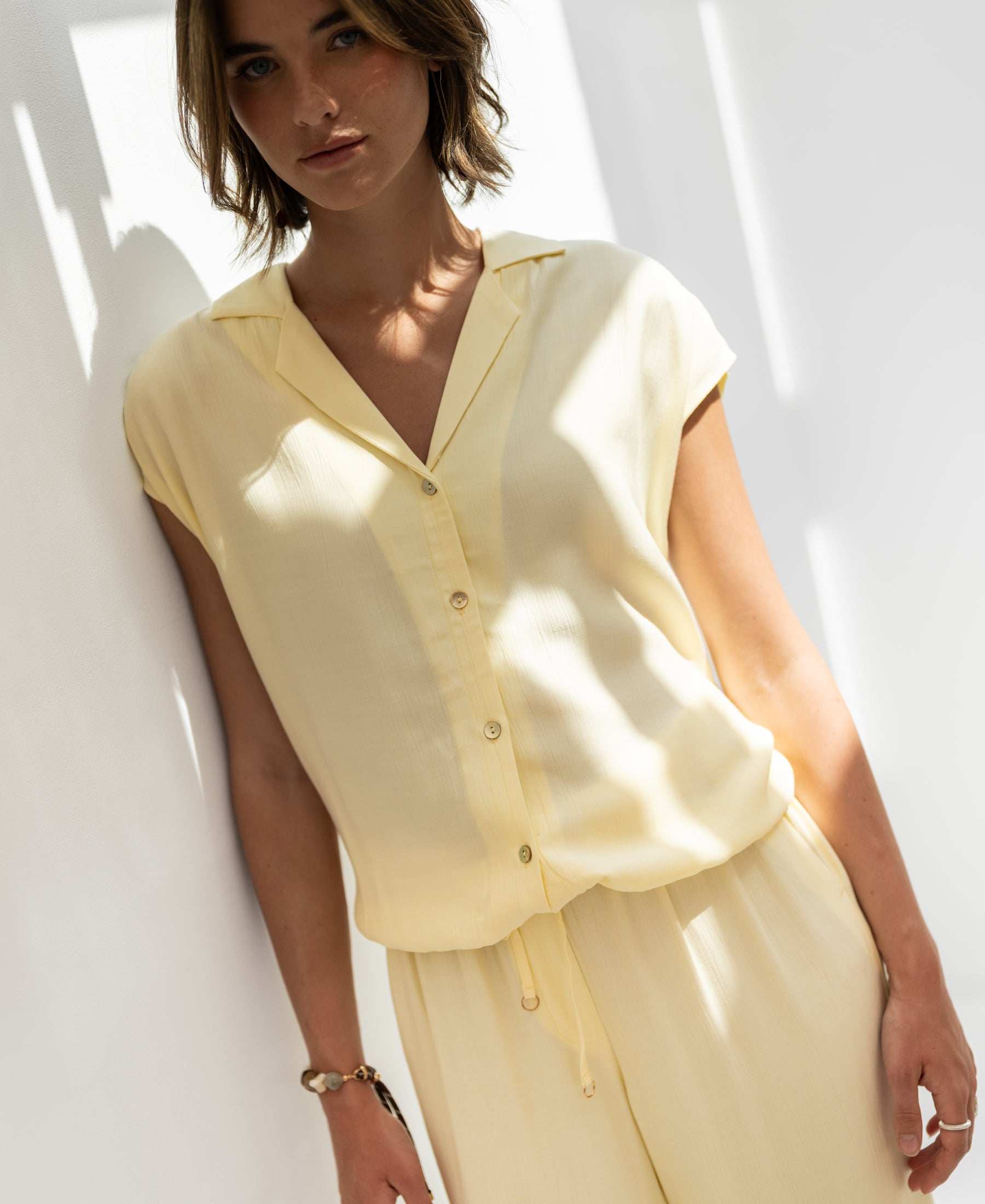 Sleeveless blouse in soft yellow by PLEIN PUBLIQUE, LA BIJOU style, worn by a woman against a sunlit white wall.