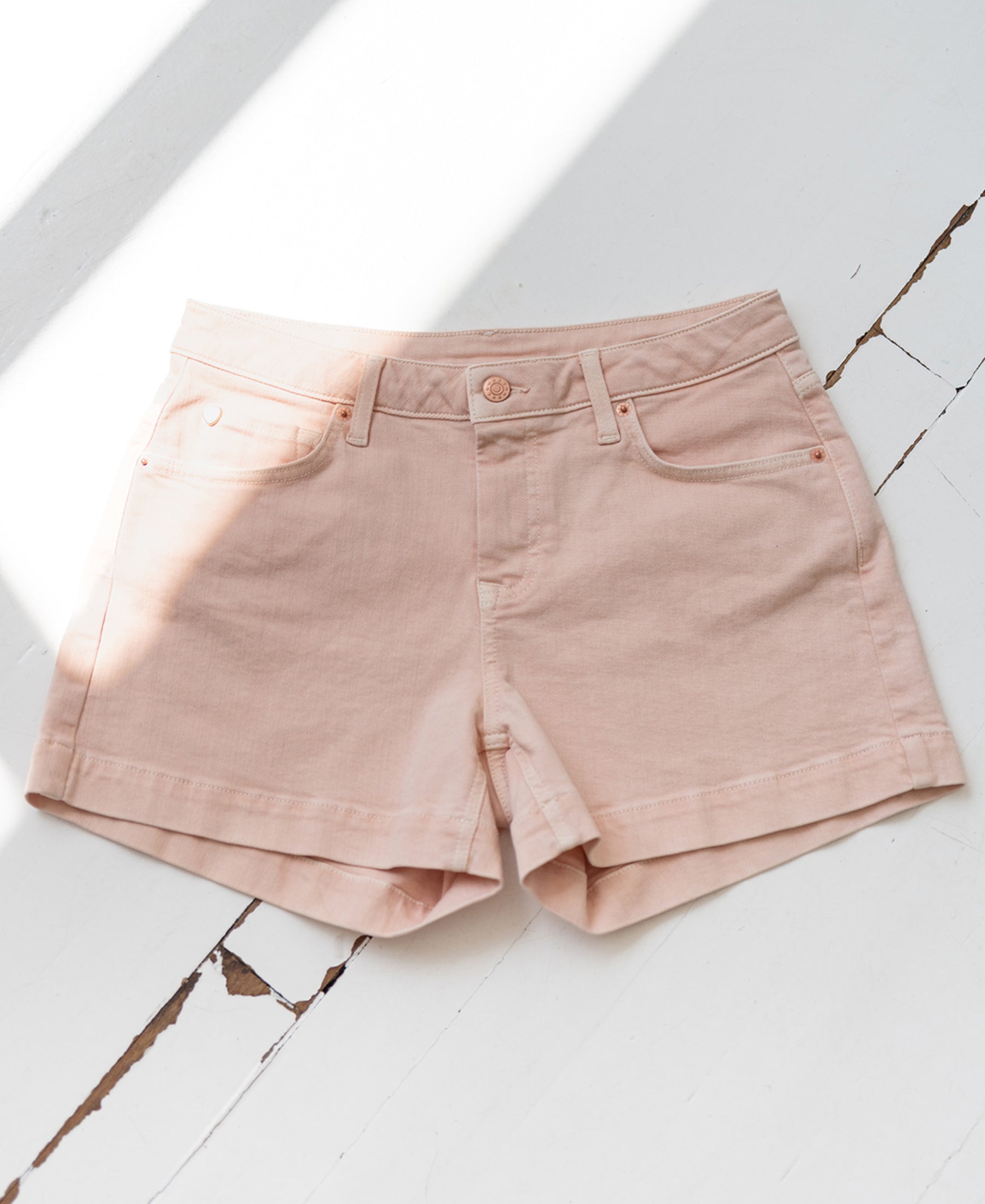 Blush pink shorts laid flat on a white, cracked wooden surface with sunlight casting across.