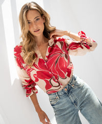 Woman wearing a ruby red print blouse, standing in sunlight with her head tilted and hand touching her hair.