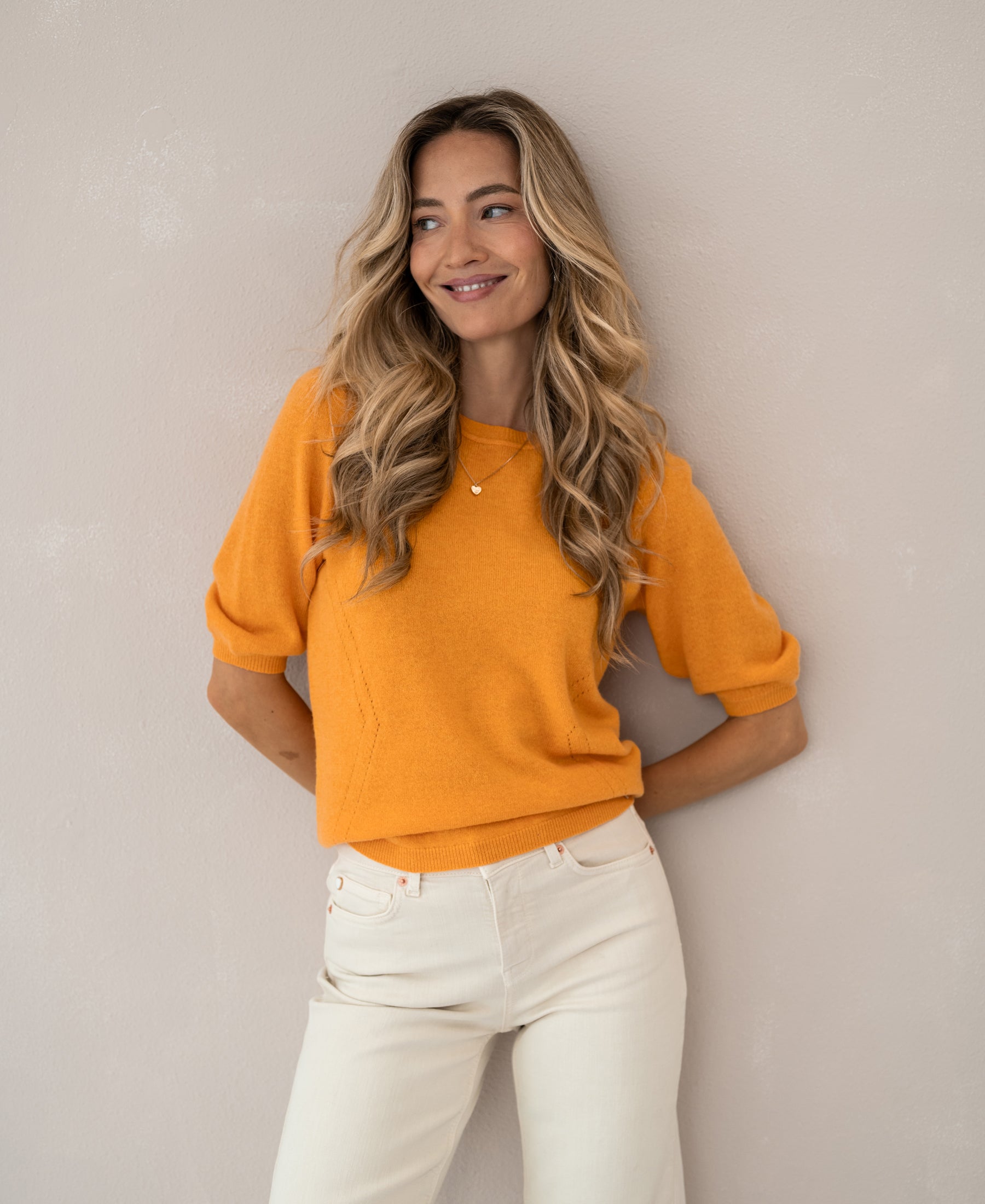 Woman wearing orange LA BLANCHE sweater stands by a light beige wall.