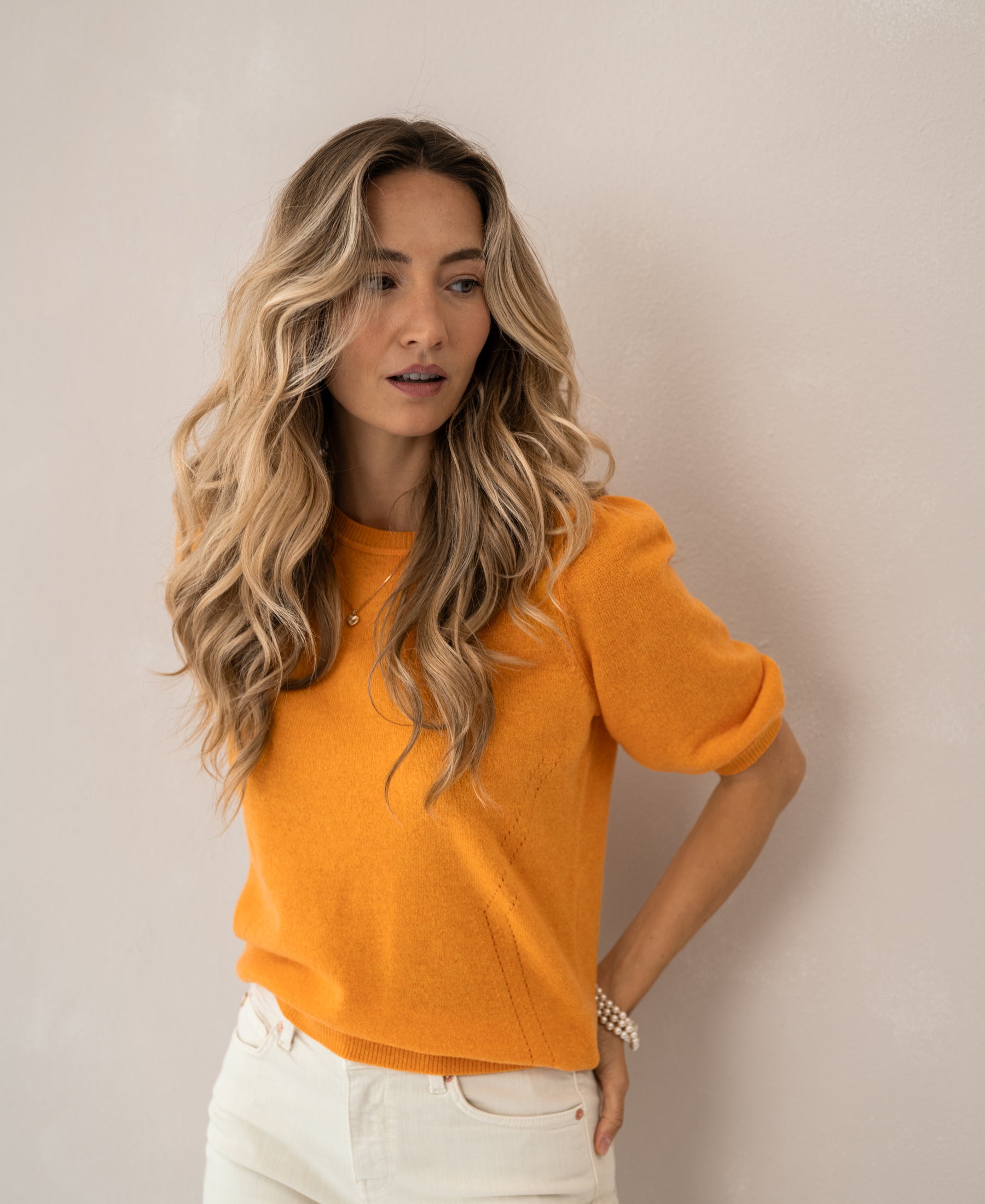 Woman wearing the PLEIN PUBLIQUE LA BLANCHE orange sweater stands against a light wall.