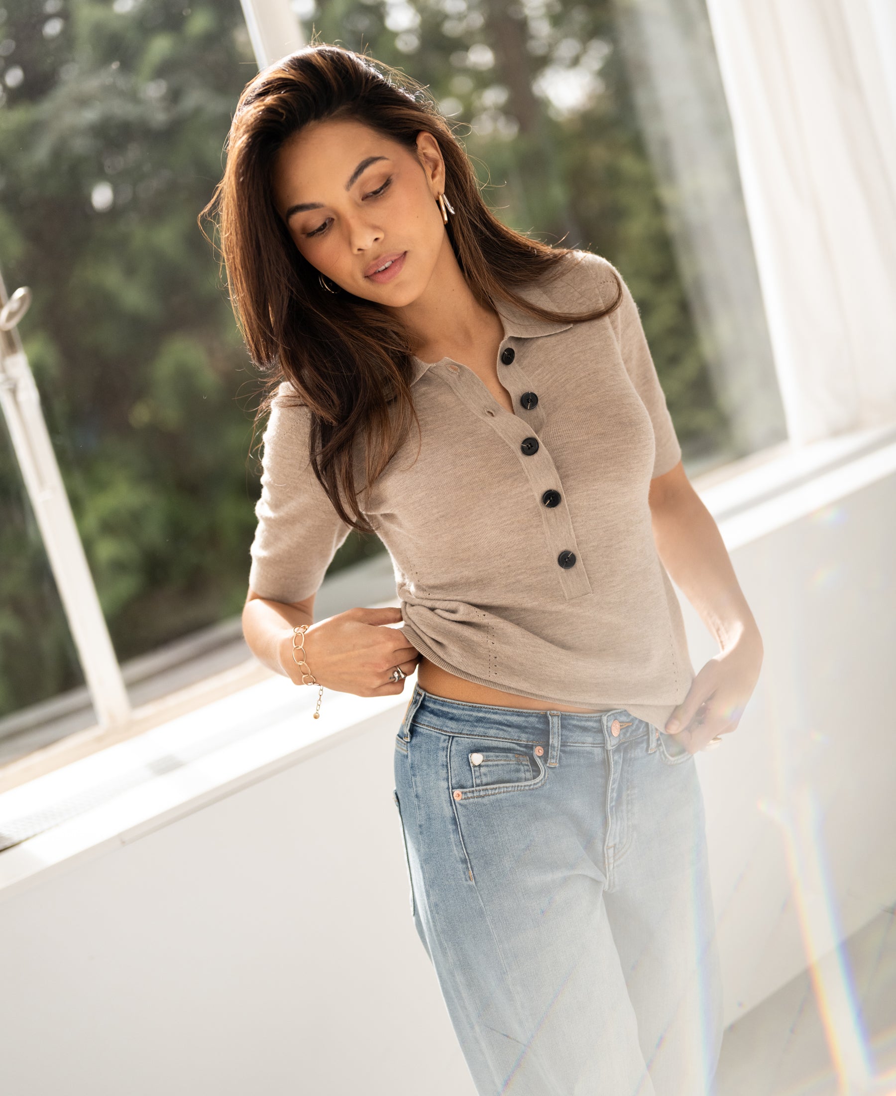 Merino wool polo shirt in dark sand by PLEIN PUBLIQUE, shown on a woman standing by a sunlit window, looking down.