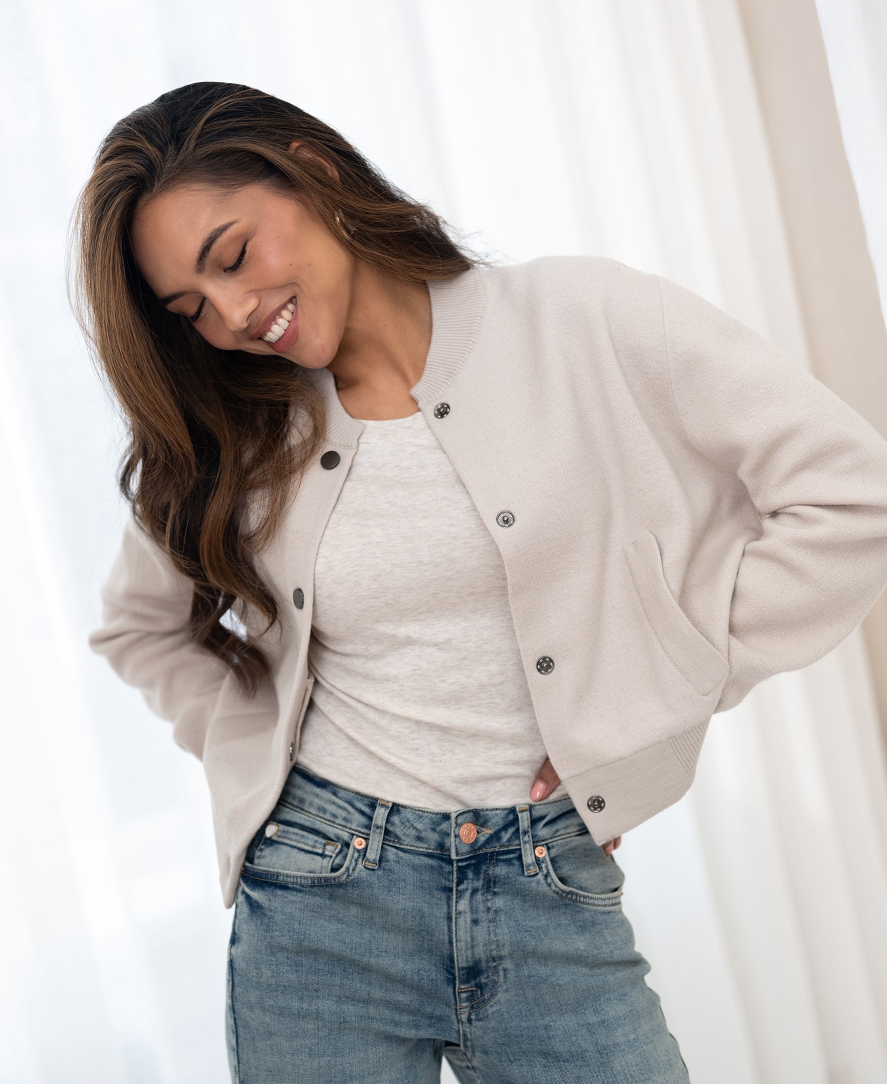 Woman wearing PLEIN PUBLIQUE's beige bomber jacket stands in front of sheer white curtains.
