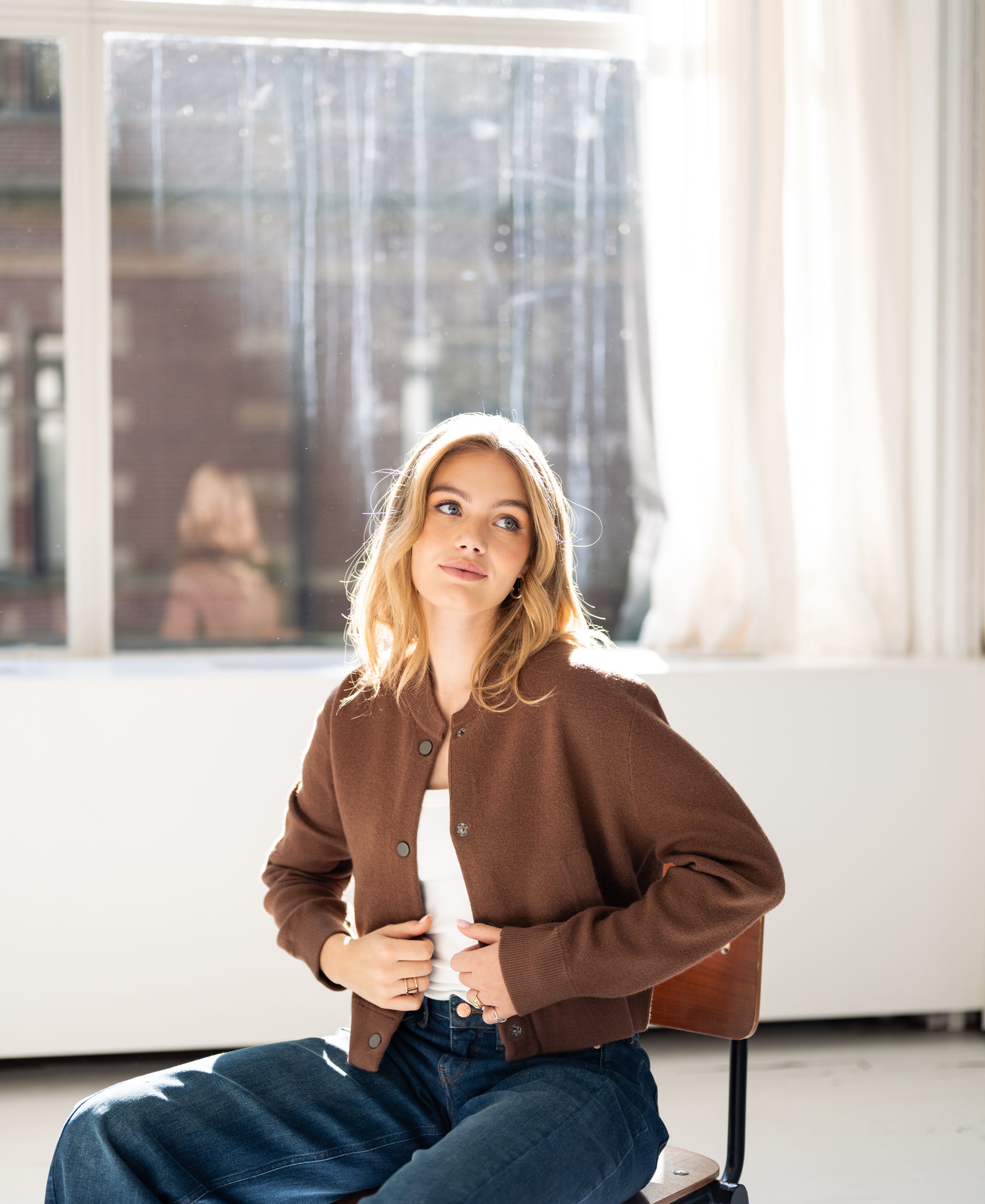 Camel bomber jacket by Plein Publique, shown on a woman sitting by a bright window, gazing sideways.