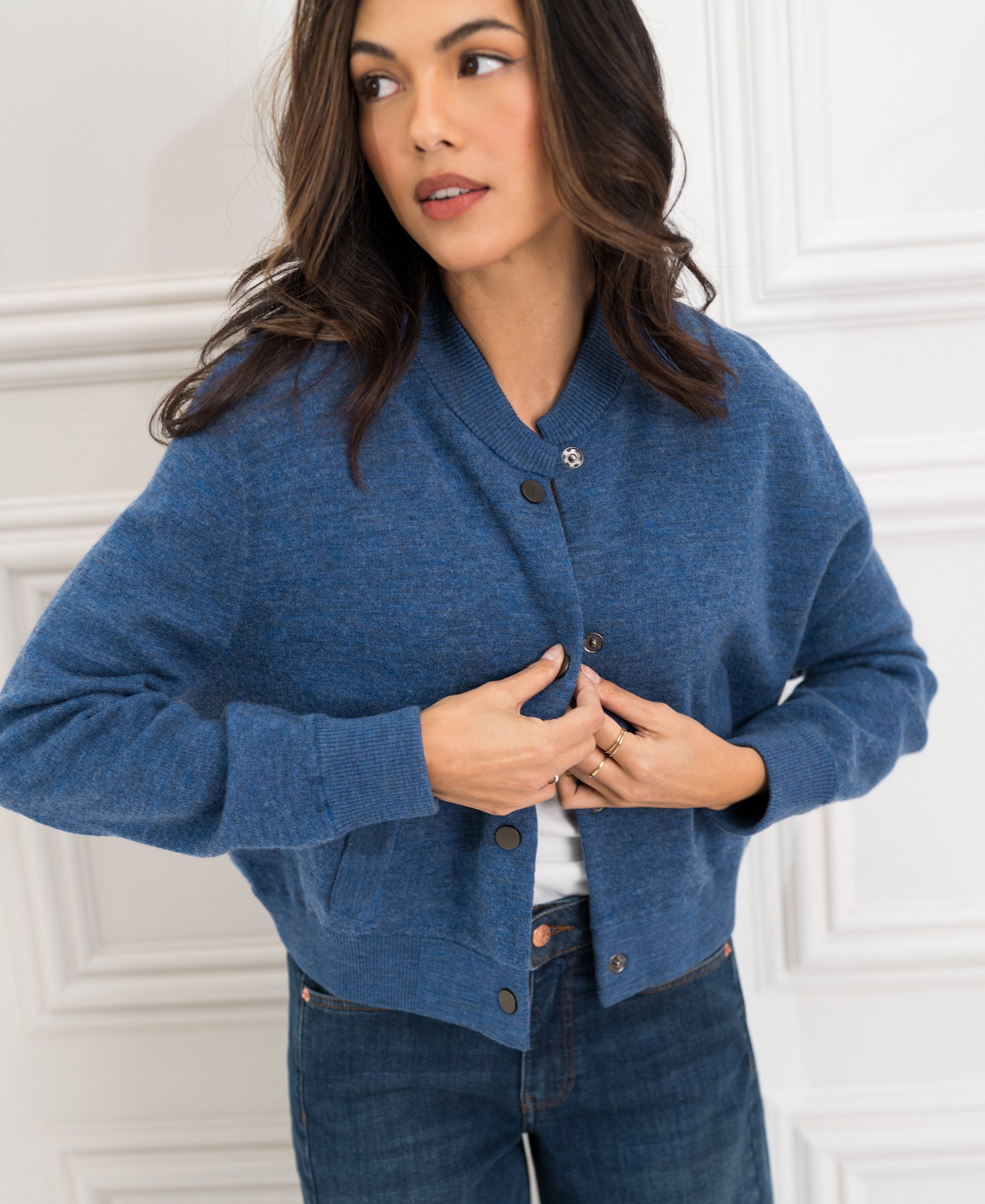 Woman buttoning up a blue denim bomber jacket by a white paneled wall.