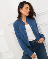 Woman wears blue denim bomber jacket by PLEIN PUBLIQUE, standing by a white paneled wall.