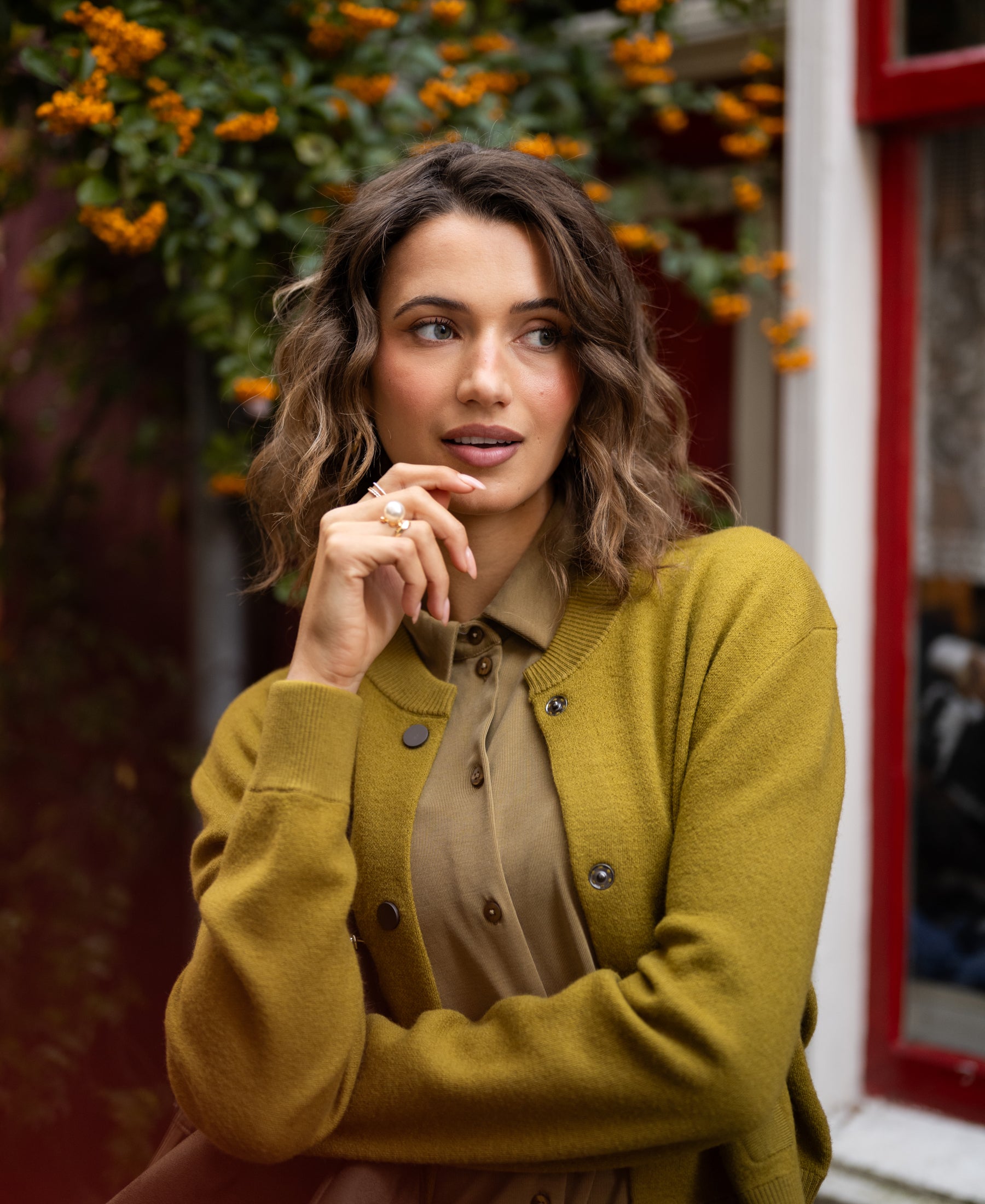 Olive green jersey blouse in a classic style shown outdoors near orange flowers and a red window.