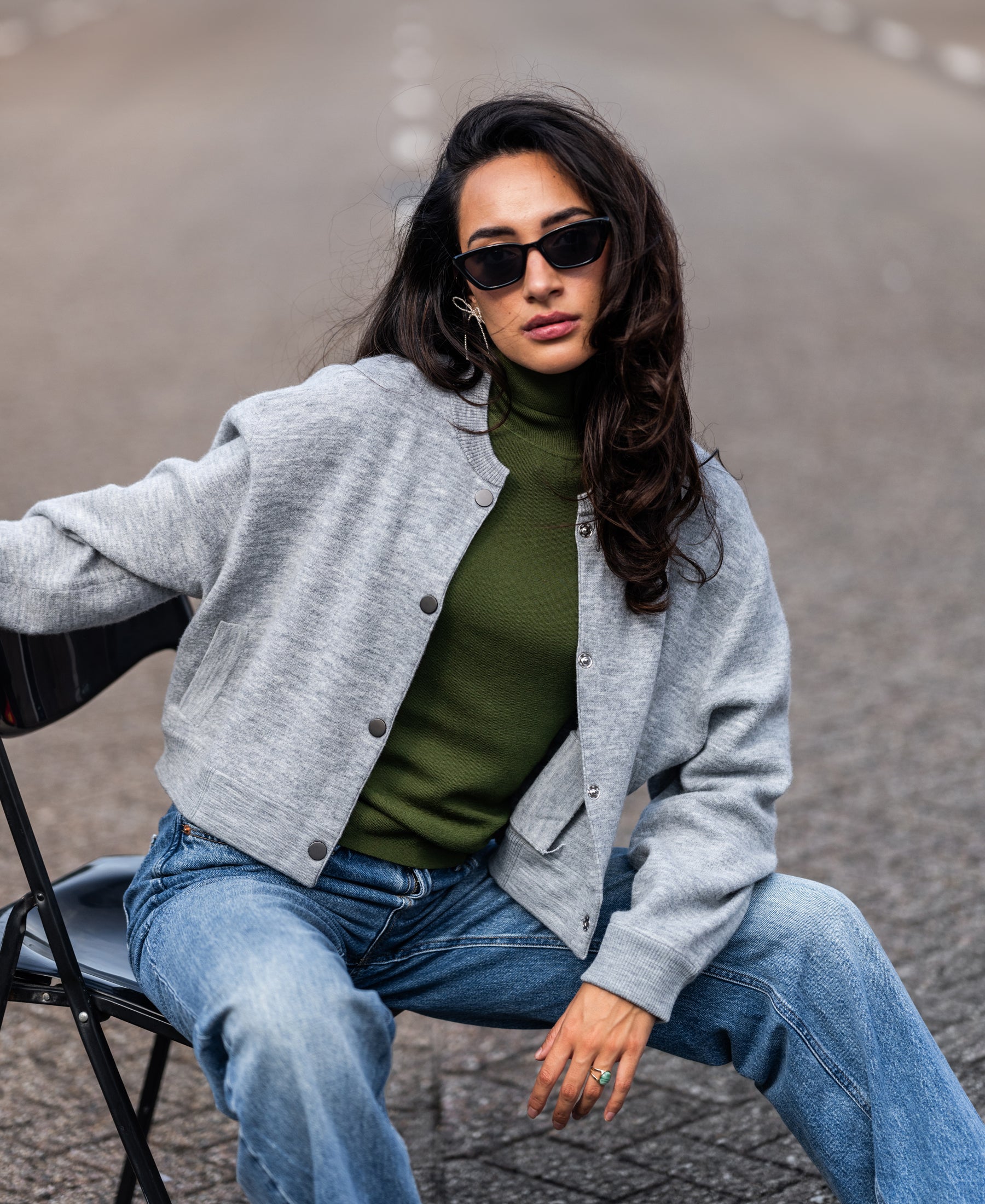 Woman wearing an olive LE FABIAN turtleneck sweater by PLEIN PUBLIQUE sits outdoors, looking at the camera.