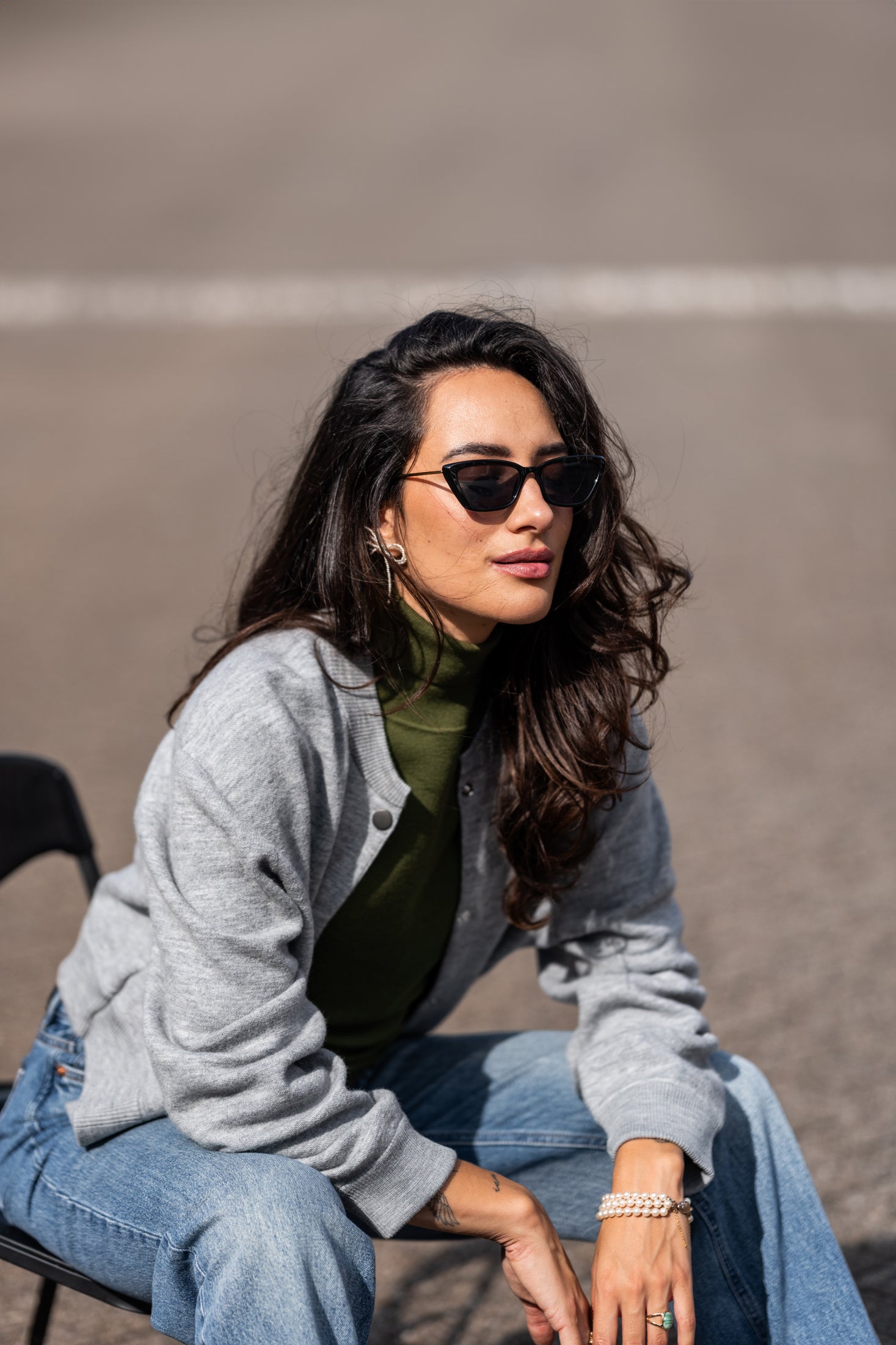 Woman wears the PLEIN PUBLIQUE LA BOMBA bomber jacket in grey melange, seated outdoors.