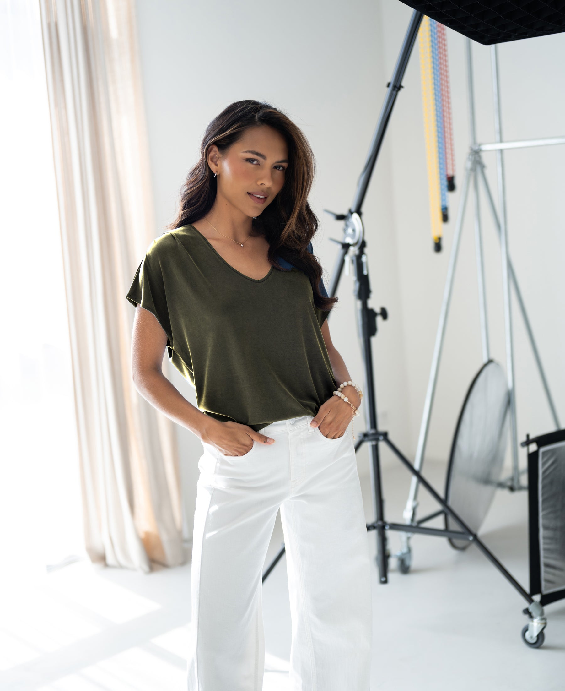 Woman wears olive green ribbed T-shirt by PLEIN PUBLIQUE in a bright studio with photography equipment in the background.