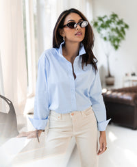 Light blue oversized blouse in cotton, shown indoors near a sunlit window.