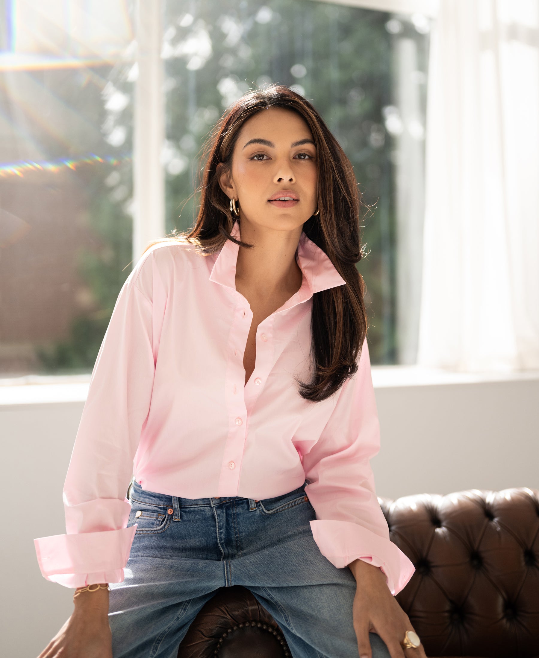 Oversized light pink LE LILLE blouse shown on a woman sitting on a brown leather sofa by large windows.