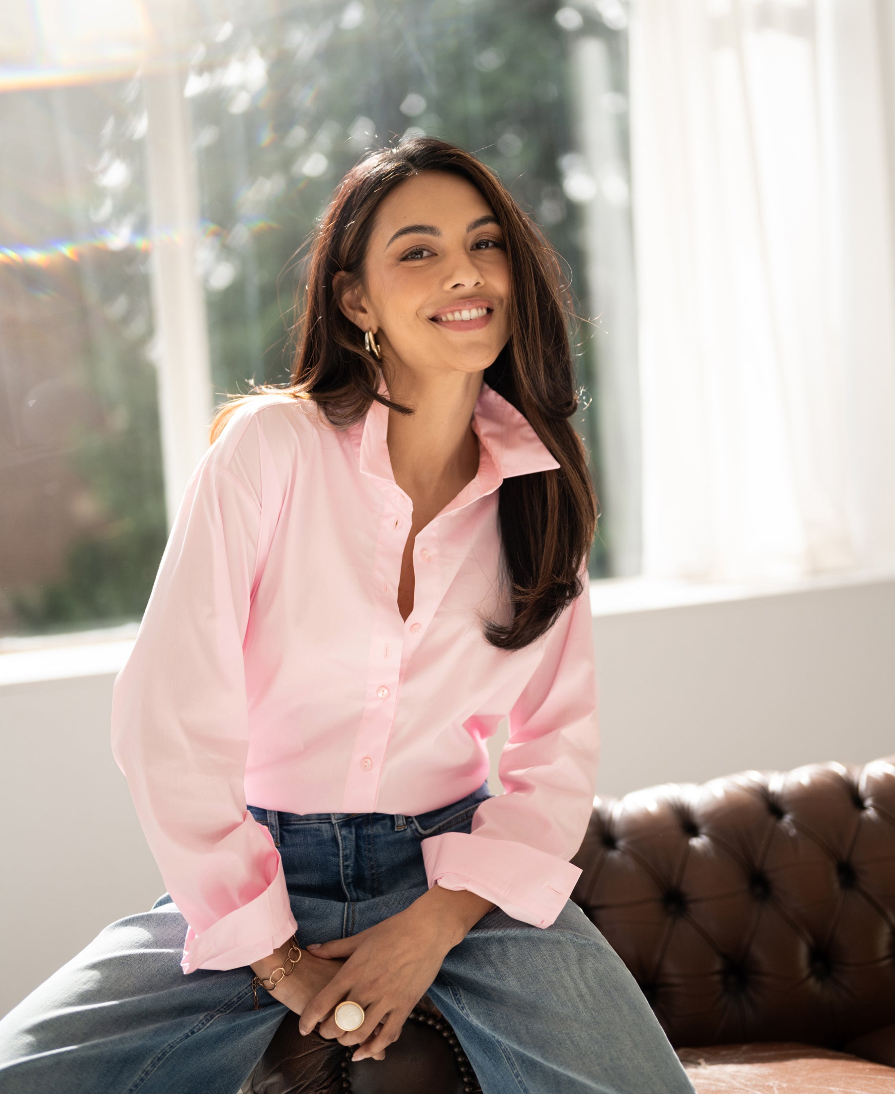 Oversized light pink LE LILLE blouse displayed on a woman sitting on a brown leather sofa near large windows.