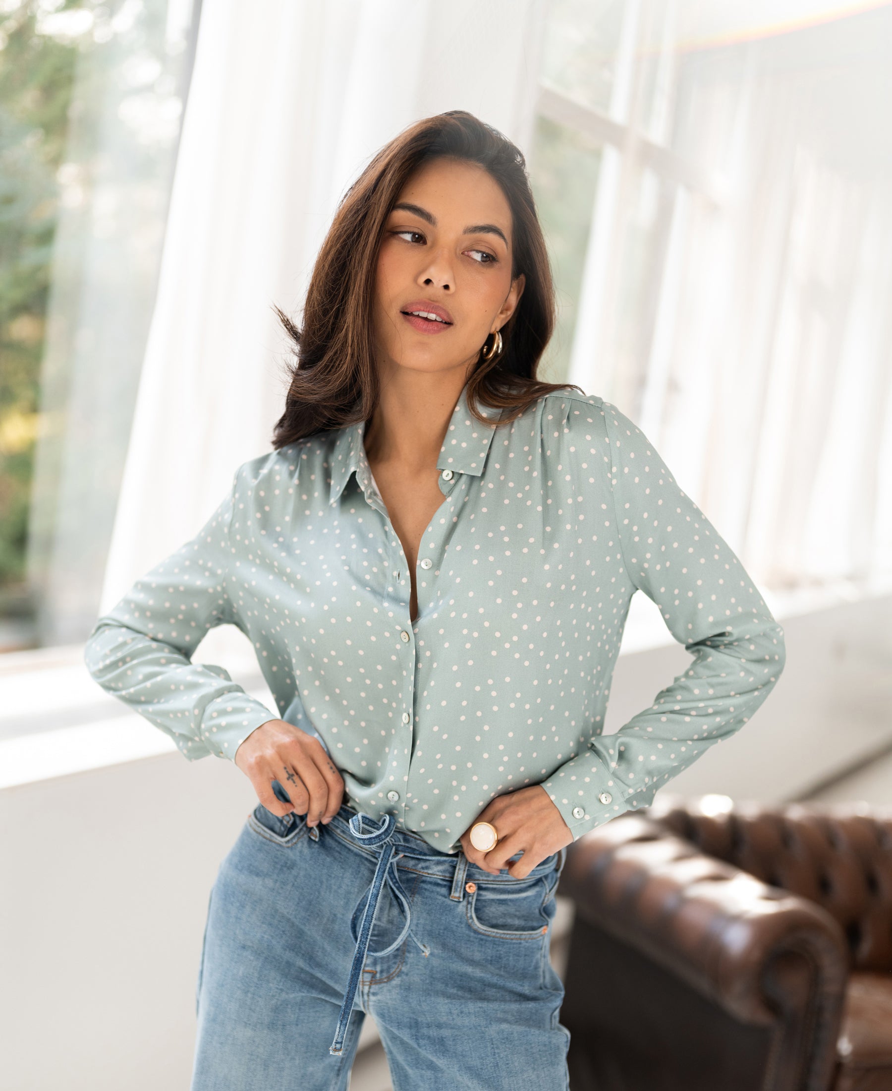 Woman wearing a light green and ivory polka dot blouse stands by a window with one hand on her hip.