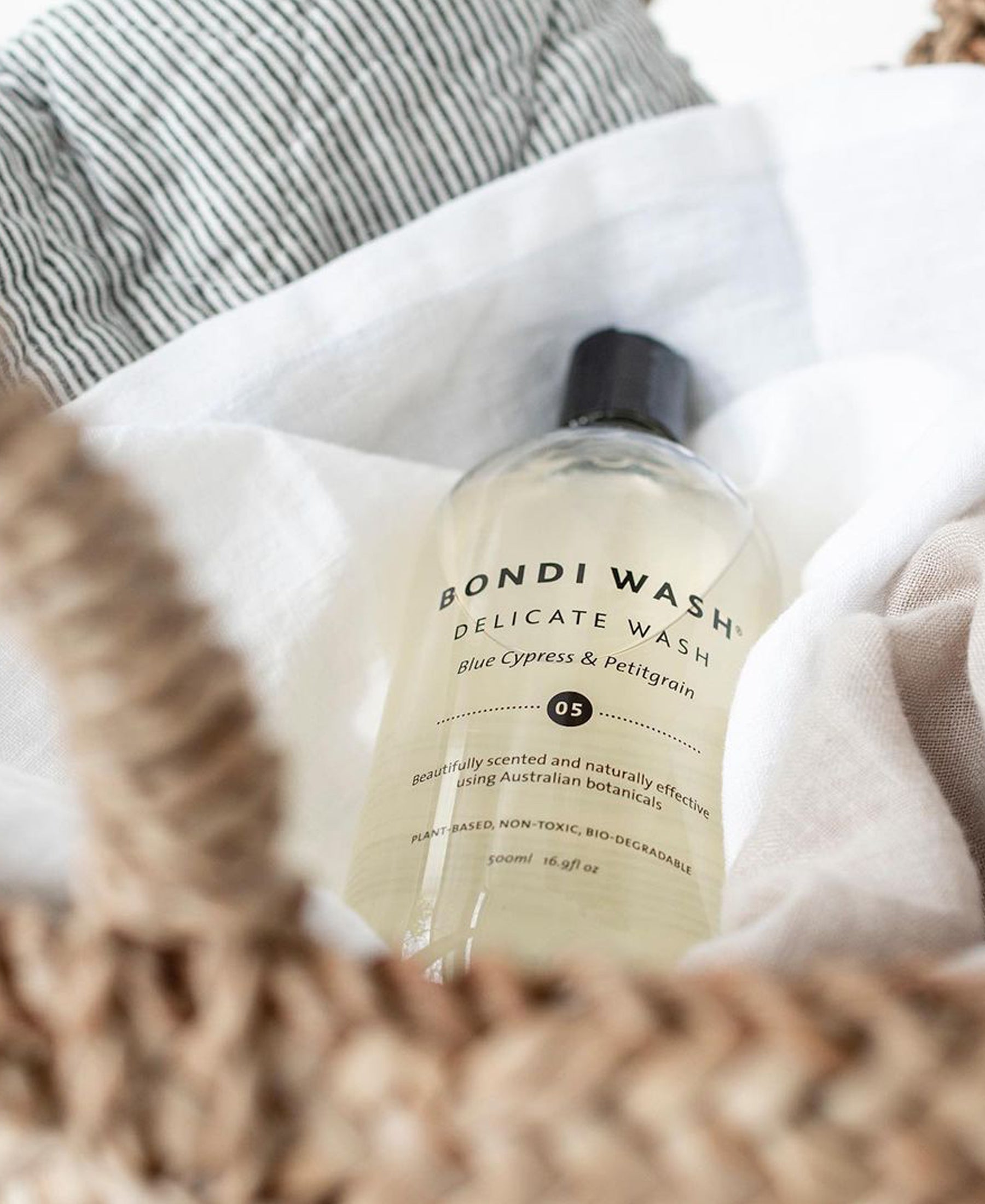 A bottle of PLEIN PUBLIQUE Delicate Wash Blue Cypress & Petitgrain on neutral fabric in a woven basket.