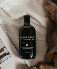 A black bottle of PLEIN PUBLIQUE Laundry Detergent BONDI WASH Fragonia & Sandalwood on beige fabric.