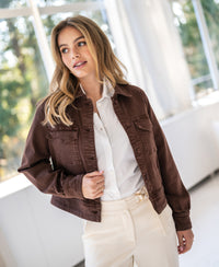 Stretch denim jacket in cacao worn by a woman with long blonde hair, standing by sunlit windows.