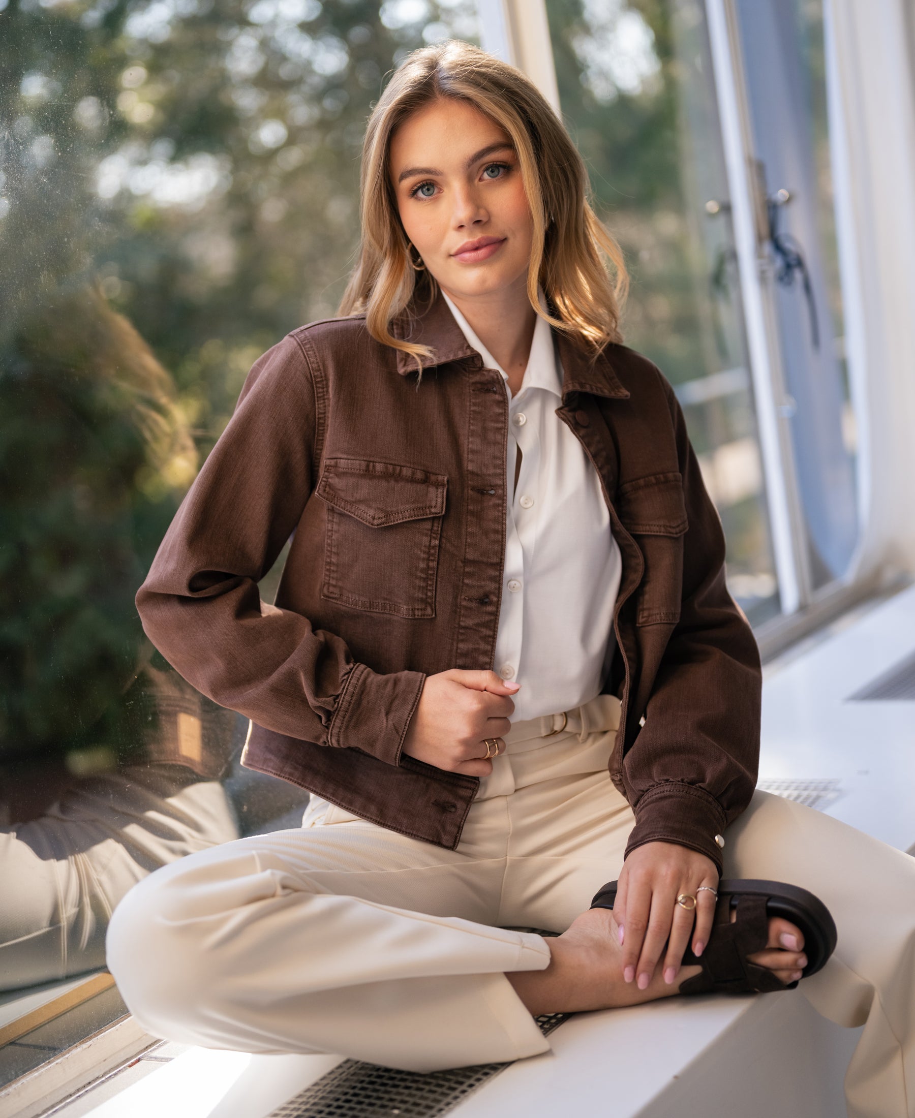 Stretch denim jacket in brown (cacao) from Plein Publique, shown on a woman seated cross-legged by a window.