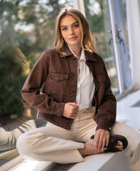 Stretch denim jacket in brown (cacao) from Plein Publique, shown on a woman seated cross-legged by a window.