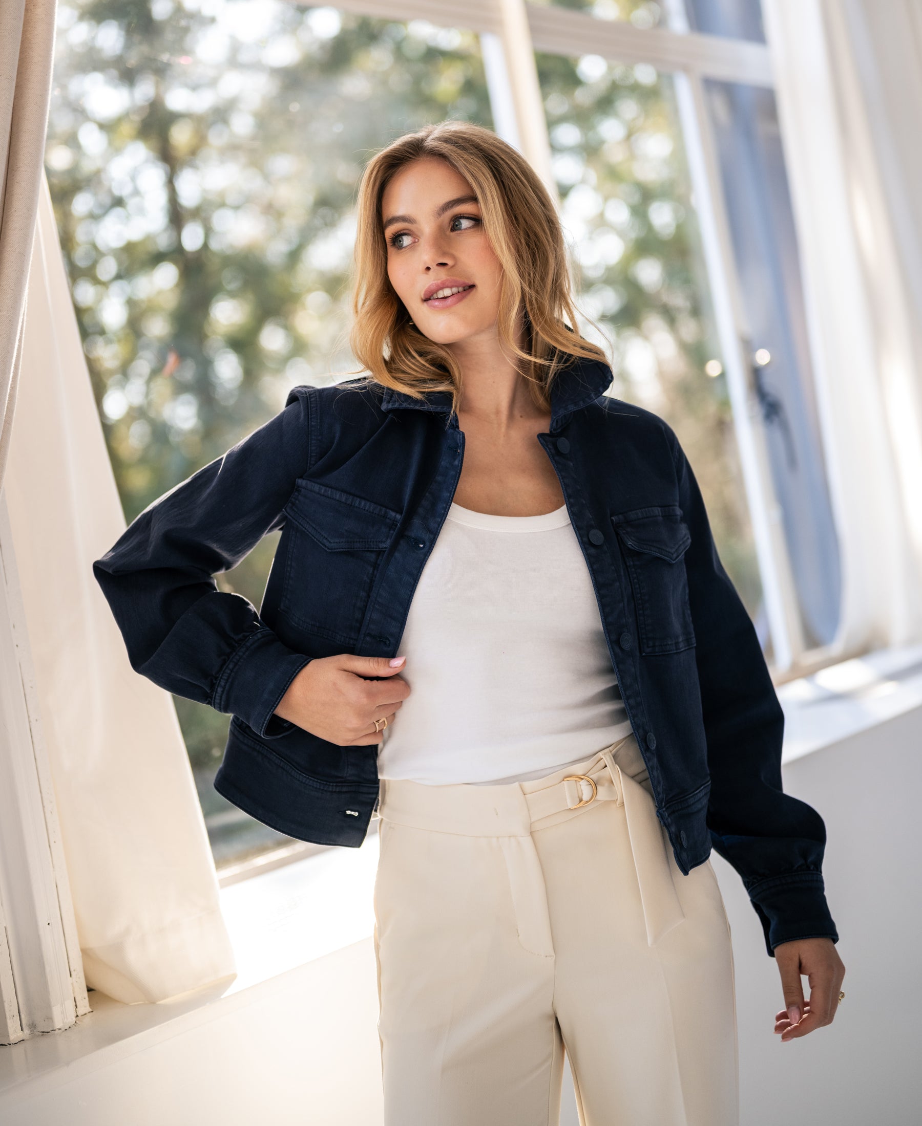 Stretch denim jacket in dark blue, shown on a model standing by a sunny window with sheer light curtains.