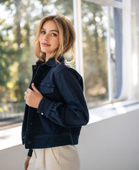 Woman wearing a PLEIN PUBLIQUE dark blue stretch denim jacket stands by a sunlit window.