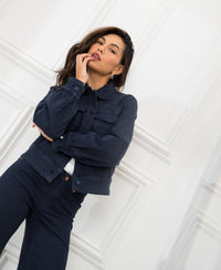 Stretch denim jacket in dark blue, shown on a woman standing by a white paneled wall.