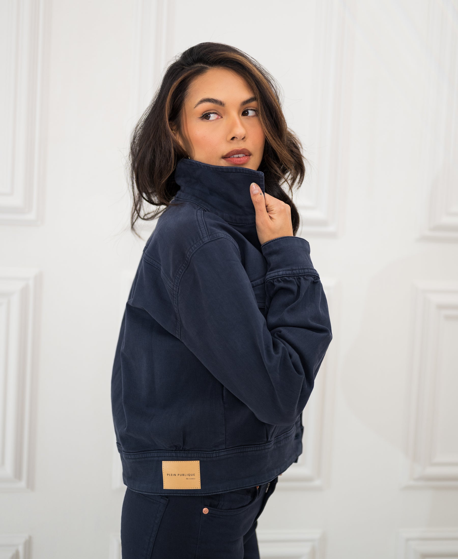 Stretch denim jacket in dark blue, shown from the back with model looking over shoulder by a white paneled wall.