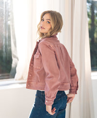 Stretch denim jacket in dusty pink, shown from the back as a woman looks over her shoulder at the camera indoors.