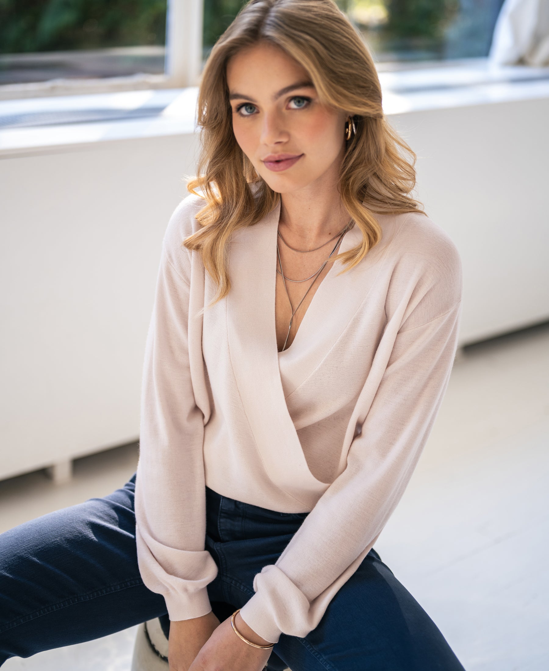 A woman wears the PLEIN PUBLIQUE LES CINQ wrap sweater in soft blush, sitting indoors and looking relaxed at the camera.