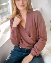 Woman wearing the LES CINQ Desert Rose wrap sweater by PLEIN PUBLIQUE sits indoors, softly smiling in sunlight.